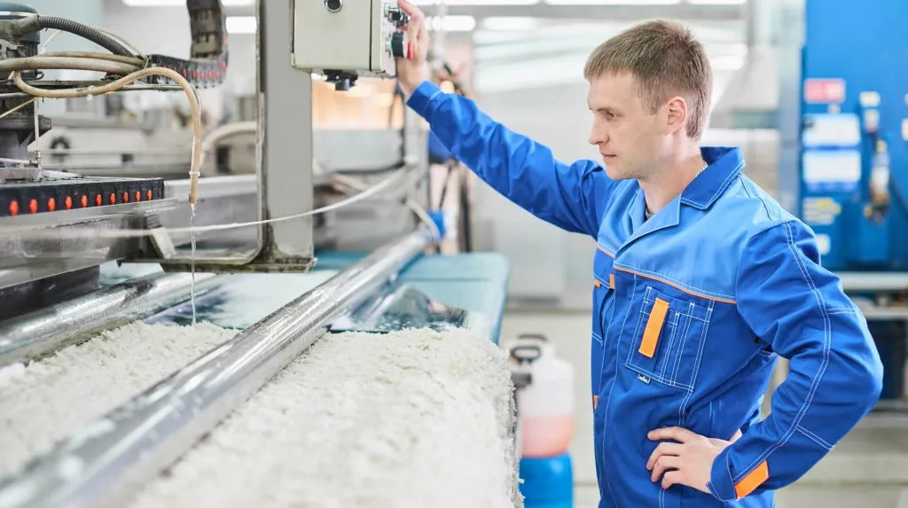 A worker in blue overalls guides a pale rug through a washing machine
