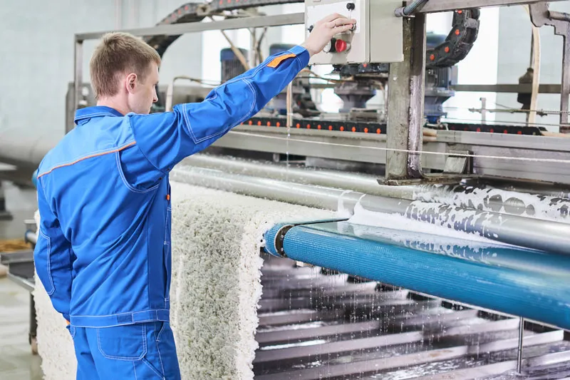 A worker in blue overalls feeds a cream rug through a washing machine at a rug cleaning facility