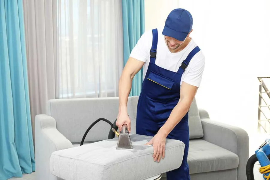 A cleaner in overalls works on a grey sofa with an upholstery tool