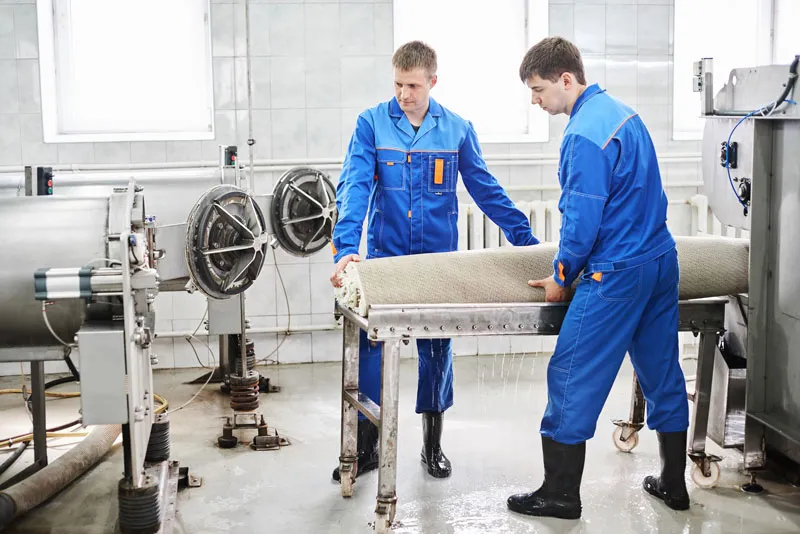 Two workers lift a rolled rug onto a cart beside large washing machines