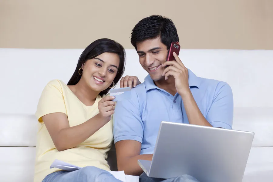 A smiling couple on a sofa, one on the phone and one with a laptop