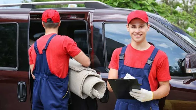 Two movers carry rolled-up rugs to their van
