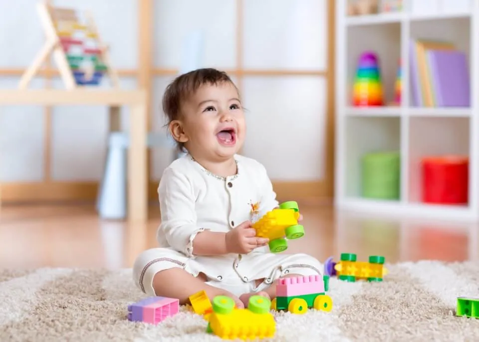 A laughing baby plays with toy blocks on a soft cream rug in a bright playroom