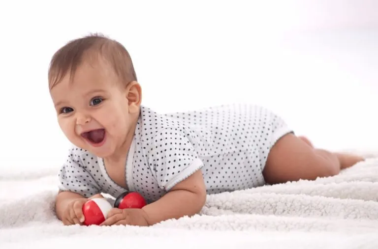 A laughing baby lies on a soft white rug, holding a toy