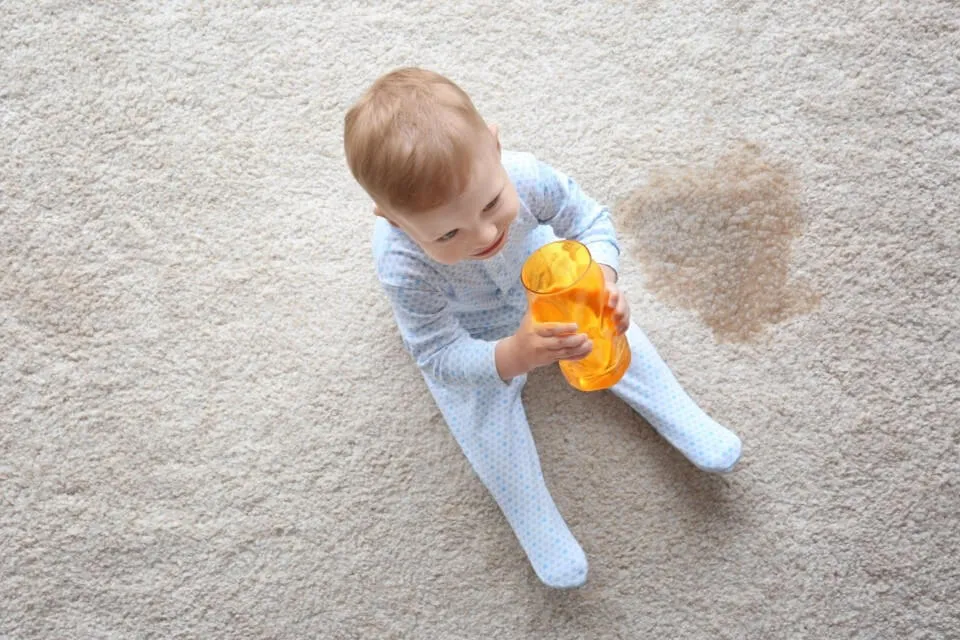 A toddler on a beige rug next to a juice stain