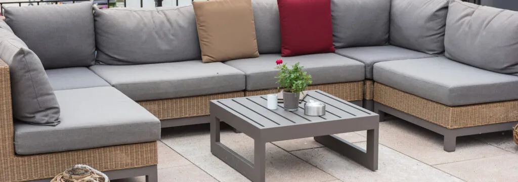 A wicker sectional with grey cushions around a low grey coffee table holding a potted rose