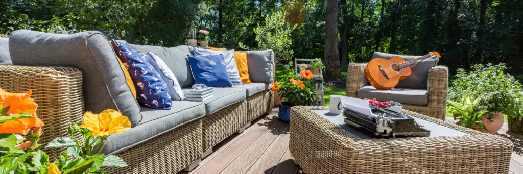 A wicker sofa with grey cushions and blue pillows on a sunny deck, a guitar resting on the armchair beside it