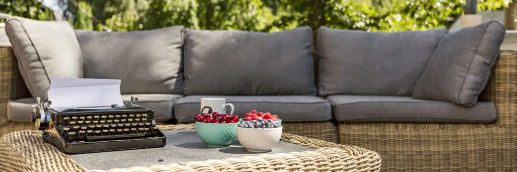 A wicker sofa with grey cushions behind a wicker table set with a typewriter and bowls of berries