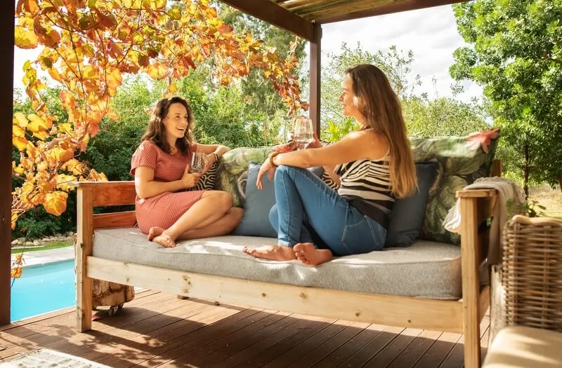 Two women chat on a cushioned wooden daybed on a covered deck by a pool, under autumn leaves