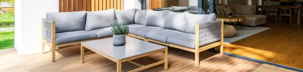 A wooden outdoor sectional with grey cushions and a low table on a covered deck outside a living room