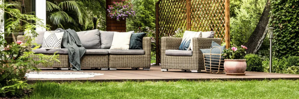 A wicker sofa and armchair with grey cushions on a deck beside a lawn and a wooden trellis