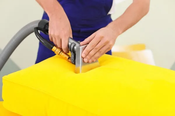 A cleaner in blue overalls runs an extraction tool over a bright yellow cushion