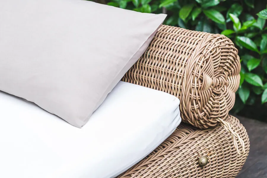 A white seat cushion and a pale pillow on a woven wicker outdoor chair in front of green leaves